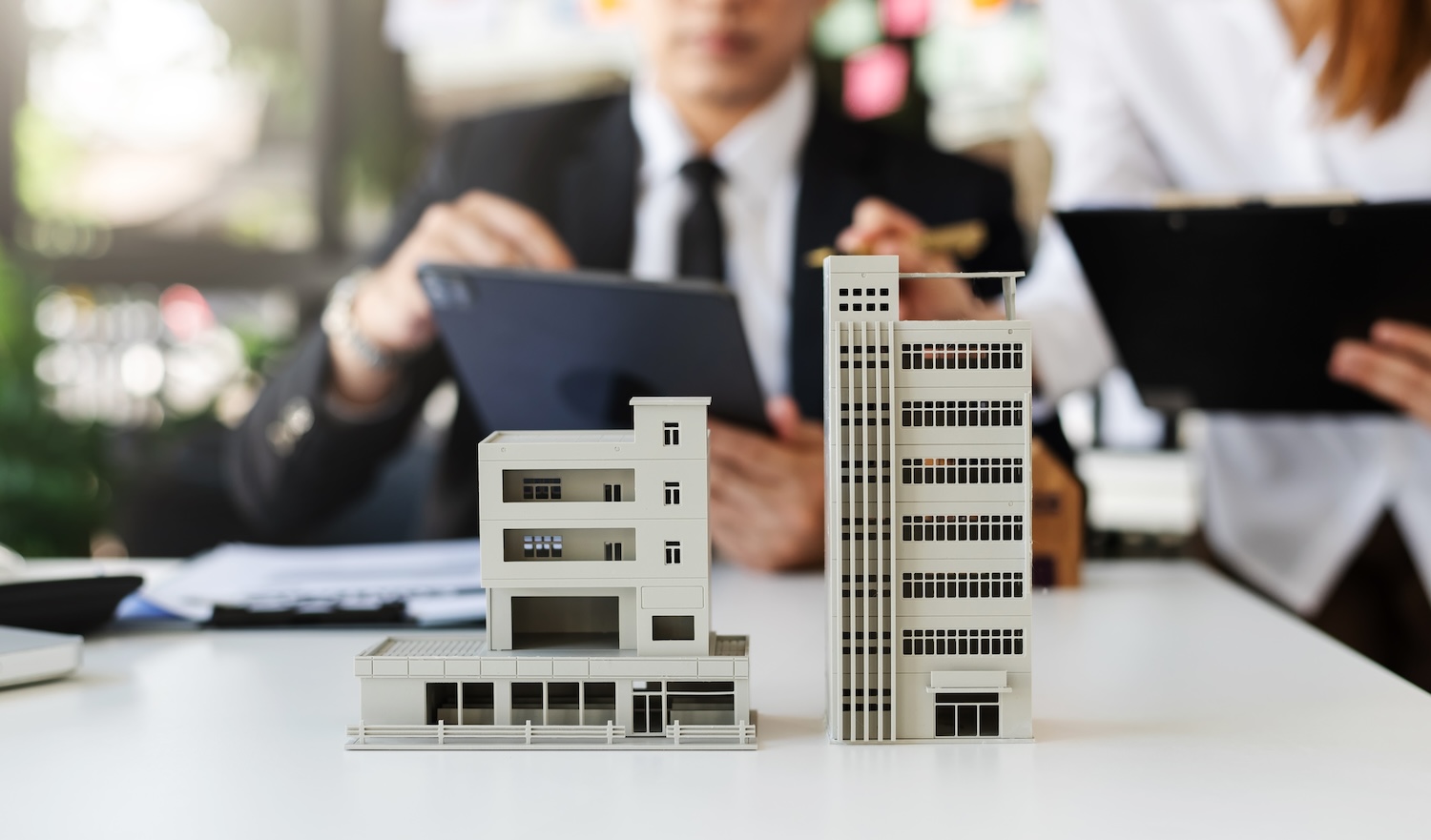 Deux modèles architecturaux de bâtiments sont posés sur un bureau blanc, avec deux individus flous en tenue professionnelle travaillant avec des documents en arrière-plan.