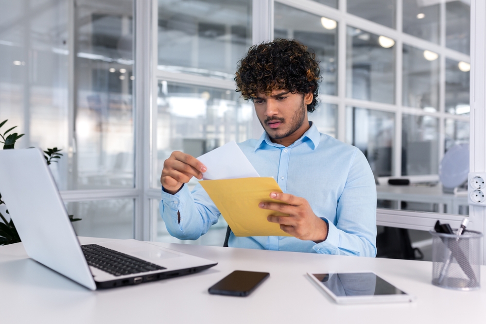 Un homme vêtu d'une chemise bleu clair est assis à un bureau, regardant un document qu'il vient de sortir d'une enveloppe jaune. Un ordinateur portable, un smartphone et une tablette sont sur le bureau.